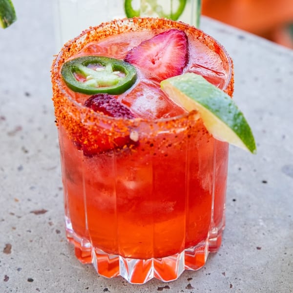 A vibrant red strawberry margarita with a chili-lime rim, garnished with jalapeño and lime.