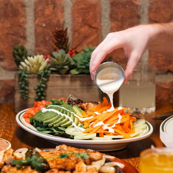 A hand drizzles dressing over a fresh La Mex salad with avocado, carrots, and cucumbers.