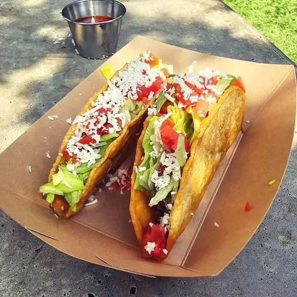 Two La Mex tacos with meat, lettuce, tomatoes, and cheese in a cardboard tray with a side of sauce.