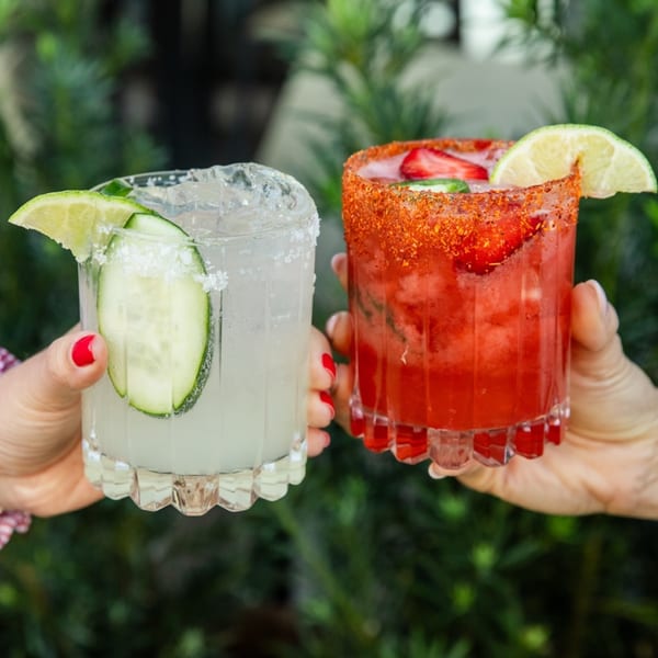 Two hands holding a cucumber margarita and a strawberry margarita with chili salt rims.