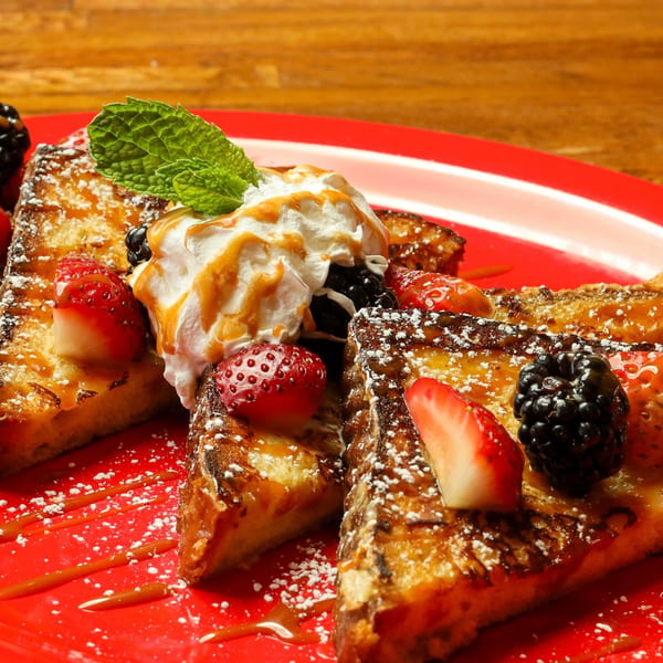 Golden French toast slices on a red plate, topped with whipped cream, berries, and caramel drizzle.