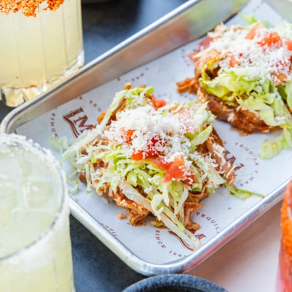 Two La Mex sopes topped with shredded meat, lettuce, tomatoes, and cheese, next to margaritas.