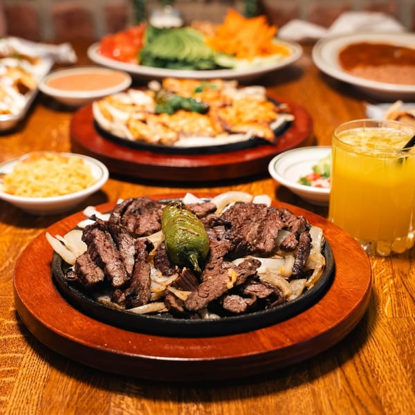 Sizzling steak fajitas with onions and a grilled jalapeño, surrounded by other Mexican dishes and a drink.