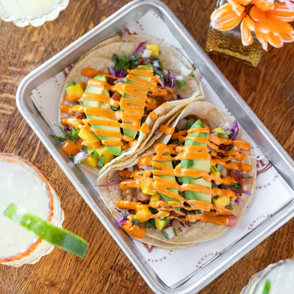 Two La Mex tacos with avocado, mango, and orange sauce, served on a metal tray with drinks.